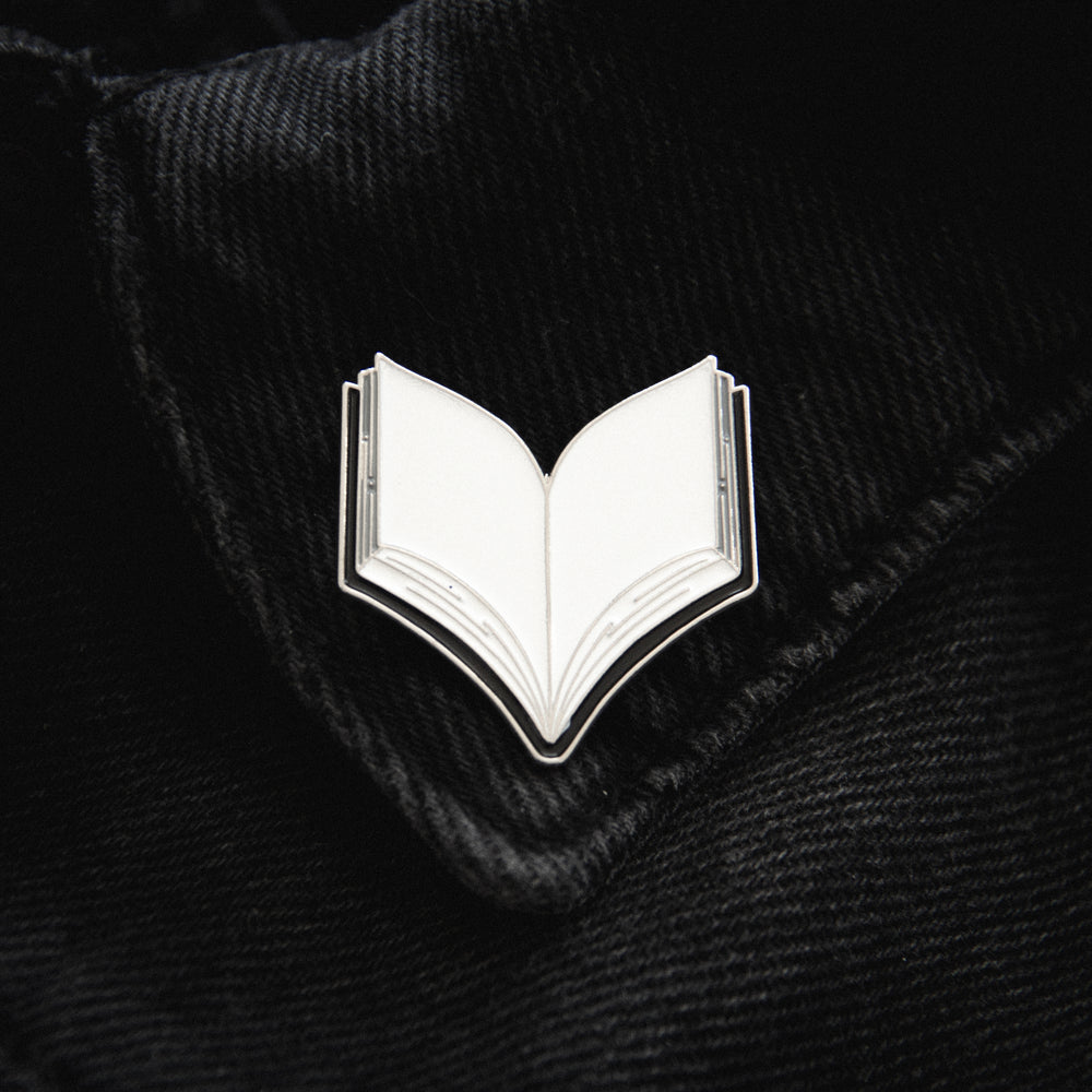 A black, silver, and white notebook journal enamel pin worn on the collar points of a shirt. 