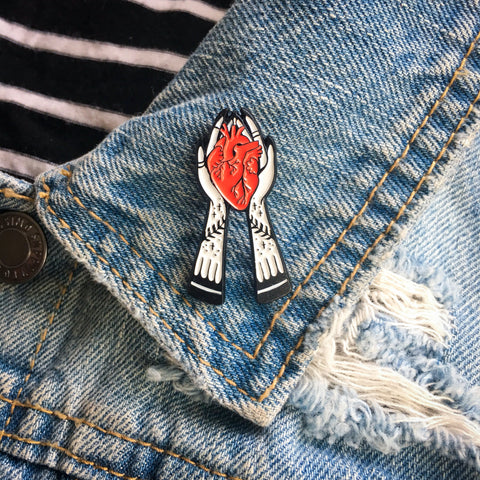 A beautiful, witchy enamel pin of a pair of tattooed hands holding a red human heart. 