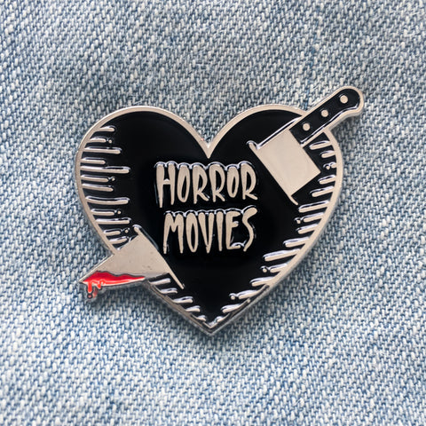 A cool enamel pin of a black and silver heart being stabbed with a bloody kitchen knife. The heart has the phrase, "Horror Movies". 