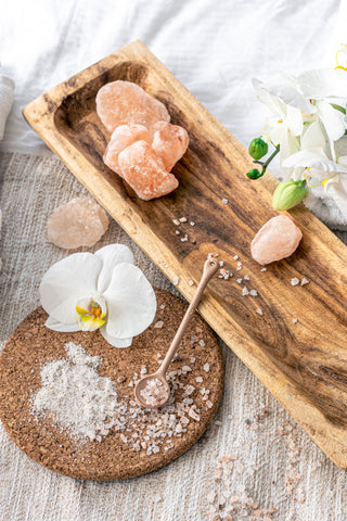 A spa composition featuring Himalayan salt chunks and granules on a wooden tray with a white orchid flower and a salt spoon on a cork mat.