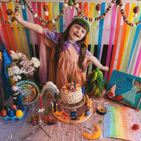 Girl with a felted crown on her head standing in front of a Waldorf birthday table with a cake, a Waldorf birthday ring with decorationsm, including a number 5