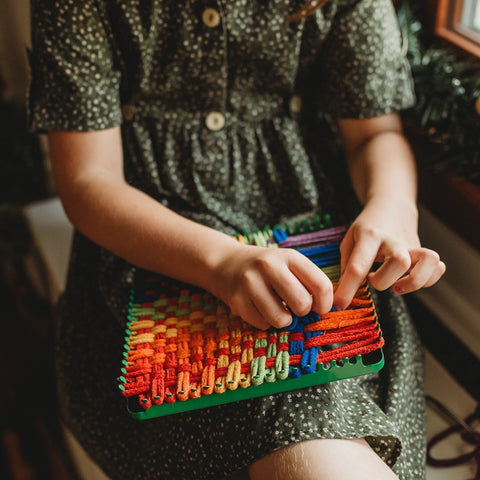 A young girl sits using a loom to weave a pot holder.