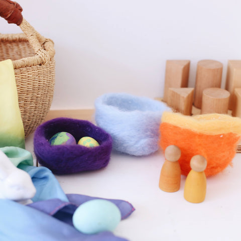 Three small felted baskets made out of wool roving in purple, light blue, and orange with a yellow stripe across the top. The purple basket holds two colored eggs