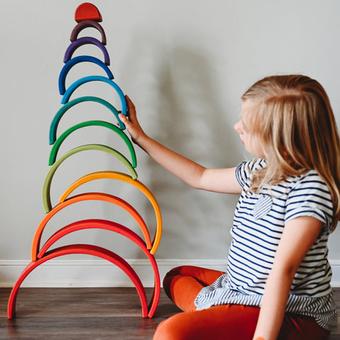 A child in a striped shirt holds her arm up to a Grimm's 12 piece rainbow stacking tunnel that she has stacked from biggest on the bottom up to smallest on top in a wavy tower