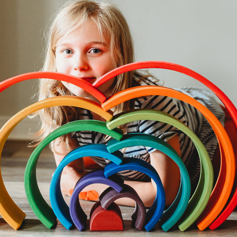 A blonde girl with a smile sits behind a Grimm's wooden toys 12 piece rainbow stacking tunnel that has been stacking in a cascading fashion