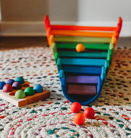 A Grimm's rainbow stacker filled upside down with their open ended sides facing up, each rainbow has a building board laid across the two side of the rainbow in cascading order from largest to smallest. A yellow bar sits on one of the building boards and three shades of red balls lay in front of it on a rug