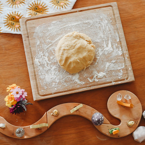 A ball of dough sits on a floured wooden cutten board on a table next to a grimm's celebration spiral with candles and ornaments, a small bouquet of flowers, and a kitchen towel with suns