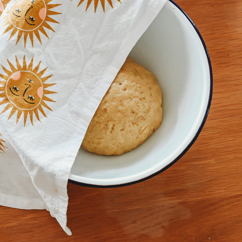 A white enamel bowl with blue rim with a large ball of dough inside covered halfway by a kitchen towel decorated with suns
