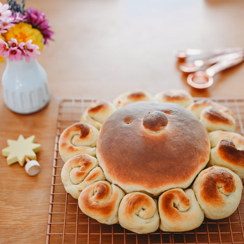 Sun bread sits on a wire cooling rack on a table next to a small white vase with yellow and purple flowers and three copper measuring spoons