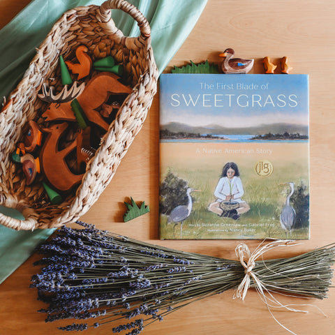 First Blade of Sweetgrass book lays on a table next to a braided grass basket with ostheimer wooden animals in it. At the bottom of the table there is a bundle of dried lavender