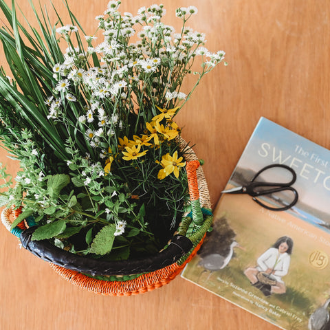 Bolga basket filled with different types of long grasses and wild flowers. Next to the basket sits the book The First Blade of Sweetgrass with a pair of pruning scissors on top