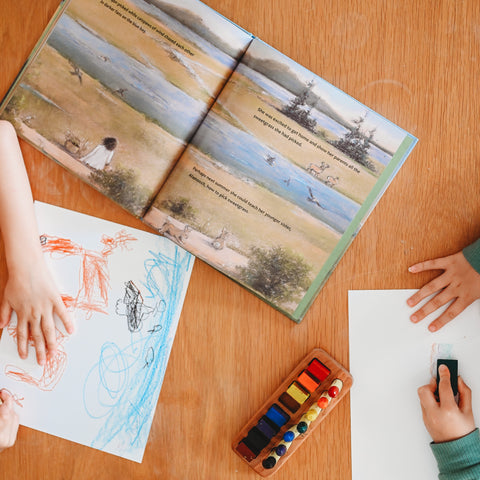 Two sheets of paper on a table being drawn on with beeswax crayons next to the book The First Blade of Sweetgrass opened to a page with an illustration of a marshy area