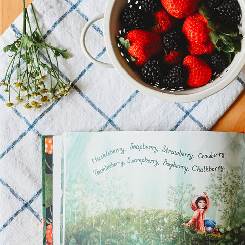 Berry Song picture book opened to berry song illustration next to a bundle of chamomile flowers and a colander with strawberries and blackberries