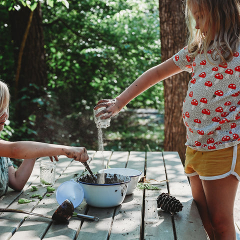 Two small children standing at a picnic table, one pours sand into an enamel bowl filled with mud while the other stirs