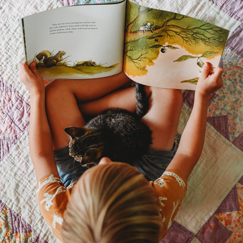 A child sitting crossed legged on a quilt with a kitten in their lap, holding the book Miss Maple's Seeds open to read