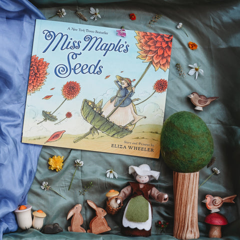 The picture book Miss Maple's Seeds laying on top of an earthy green playsilk and a blue playsilk. Surrounding the books are small flowers and seeds, a wood and wool felt tree, wooden animals such as bunnies and birds and a felted dollhouse doll dressed in an apron and brown dress