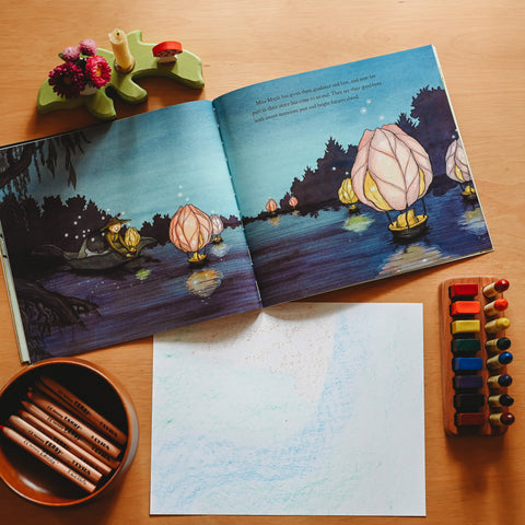 An overhead view of the picture book Miss Maples Seeds opened, sitting with a piece of drawing paper, a wooden bowl with lyra ferby colored pencils and beeswax crayons in a wooden holder