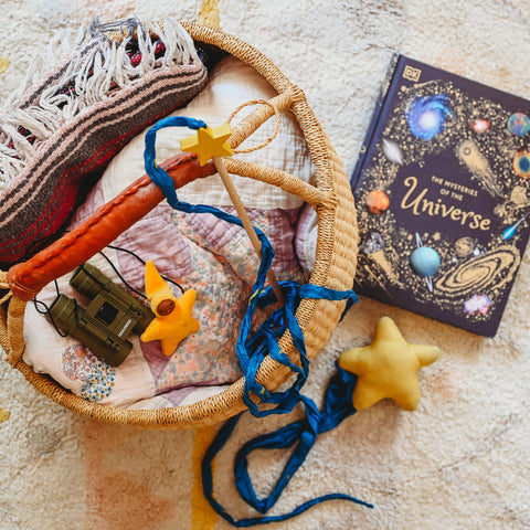 Blankets, binoculars and starry silk toys packed in a bolga basket next to a book called "Mysteries of the Universe"