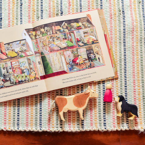 Picture book Farmhouse opened to a page illustrating the inside of a farmhouse on a braided rug next to an ostheimer wooden cow and dog and a grapat nin peg person