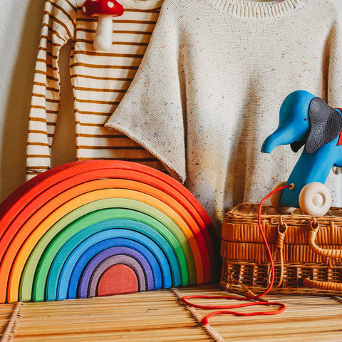 Wooden 12 piece rainbow stacking arches toy from Grimm's on a table with a wicker suitcase and blue wooden elephant pull toy waldorf from Bella Luna Toys