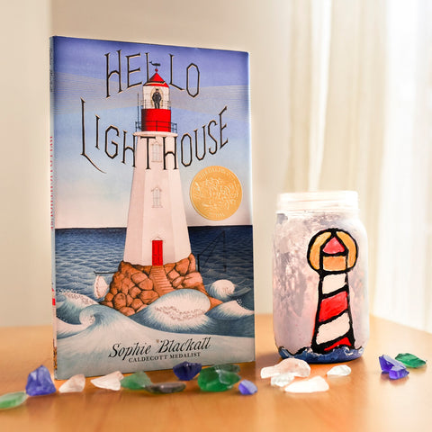 The children's book Hello Lighthouse stands on a table with a few crystals scattered in front of it. To it's right there is a homemade lantern made with a recycled glass jar to look as if it is stained glass