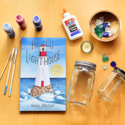 The book Hello Lighthouse lays on a table surrounded by three bottles of acrylic paint, paint brushes, school glue, two glass jars, a tea light candle, and a small bowl of blue, green, and clear cystals