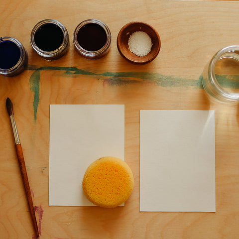 Two small sheets of water color paper on a painting tray with a natural sponge on top of one. Three jars of watercolor paints and a small bowl of salt sit to the left of the paper with a paintbrush