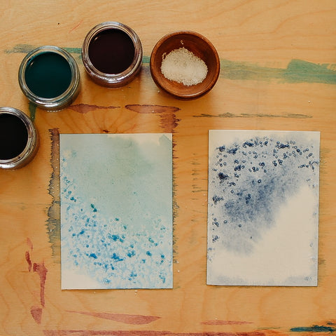 Watercolor paint in glass jars and two sheets of paper painted in greens and blues with salt sprinkled across