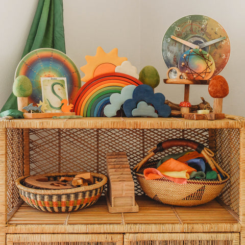 A rattan shelf with low bolga baskets on the bottom shelf and multiple waldorf toys and learning materials on the top shelf. A green playsilk hangs in the background