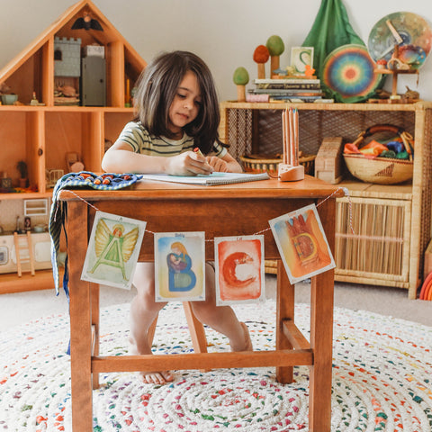 A child with dark, long hair sits at a wooden table coloring with a beeswax crayon. Hanging on the front of the table are waldorf letter cards of the letters A-D. On the table next to the paper there is a crayon roll