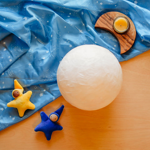 Moon lantern made out of paper mache on top of a starry playsilk next to a moon shaped tea light candle holder and two star baby waldorf dolls from Bella Luna Toys