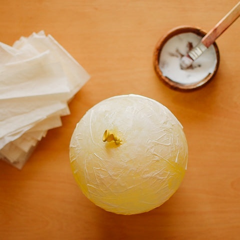 A yellow balloon covered with glue and layers of tissue paper next to a bowl of white glue with a paint brush in it and a pile of white tissue paper