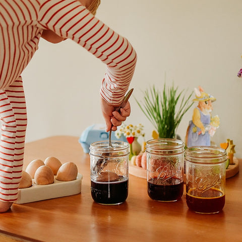 Child in white and red striped pajamas puts a slotted spoon into a glass jar with dark purple colored liquid and a wooden egg in it. Behind the jars of coloring stands a paper rabbit doll and a growth of wheatgrass