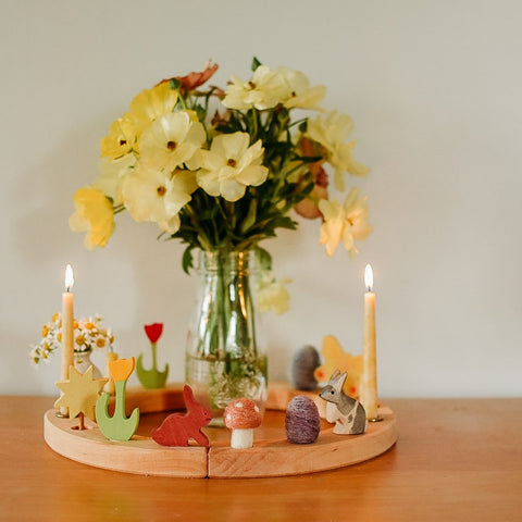 Vase of pastel yellow flowers in glass bottle with grimm's 16 hole birthday ring around it. Birthday ring has two lit candles and spring decorations like rabbits, needle felted eggs, a pink felt mushroom, a yellow tulip ornament...