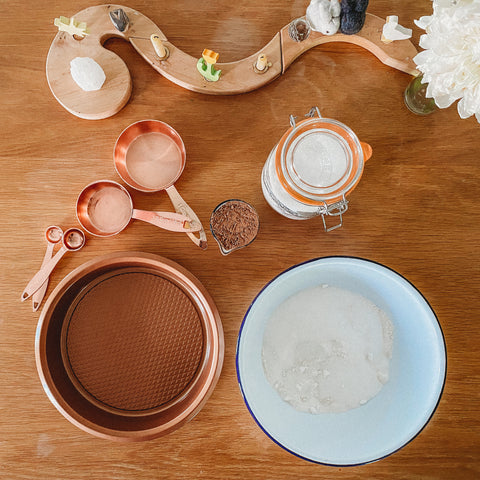 Brass measuring cups, enamel bowl filled with flour and sugar, cake pan and glass jar of sugar on a table next to a Grimm's celebration ring