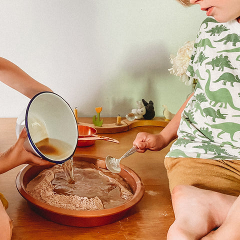 Close up image of two white children sitting on a table, pouring wet ingredients into a cake pan full of dry ingredients