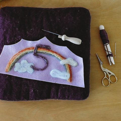 A light purple felt waldorf birthday crown with a pastel colored rainbow, clouds and deep purple number 5 laying on top of a felting mat, laying next to the mat is embroidery thread and small golden embroidery scissors