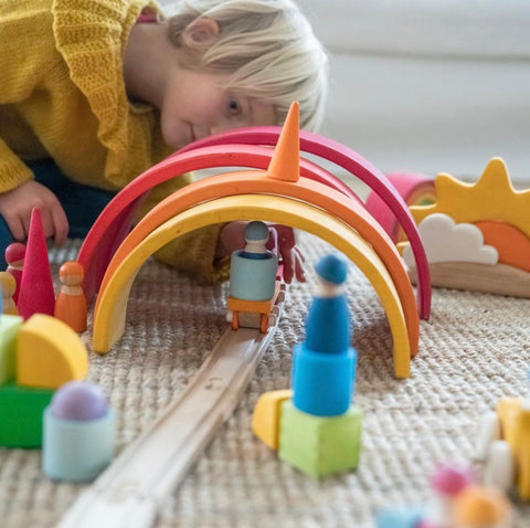 Four grimm's rainbow stackers made into a tunnel going over a wooden train track with a young girl behind it pushing a grimm's train underl the tunnel