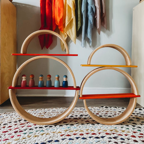 A Grimm's natural rainbow stacker stacked into large circular shapes with rainbow building boards in between them and peg friends on the boards