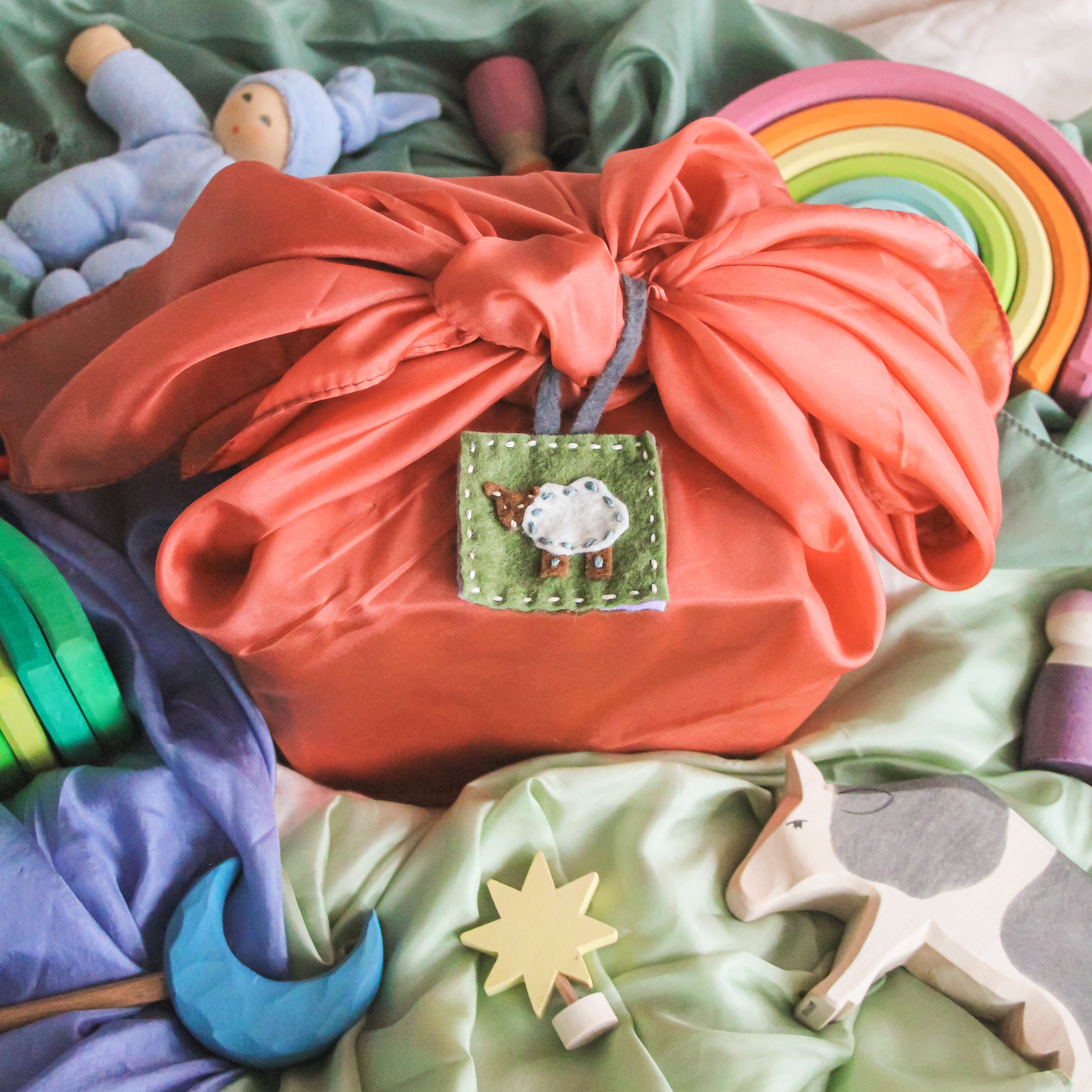 A felted reusable gift tag sits on a playsilk wrapped gift, with Waldorf inspired toys surrounding it.