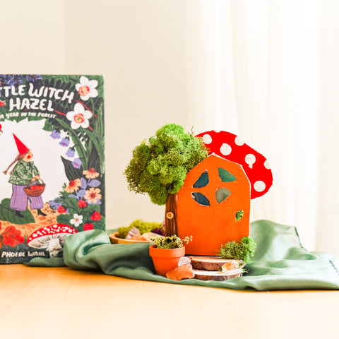 Handmade fairy door created with cardboard, paint and moss on a table sitting on a green playsilk with the book Little Witch Hazel to the left 