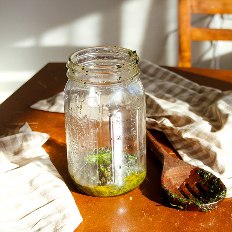 A mason jar sits with mixed mint and olive oil with a wooden spoon next to it.