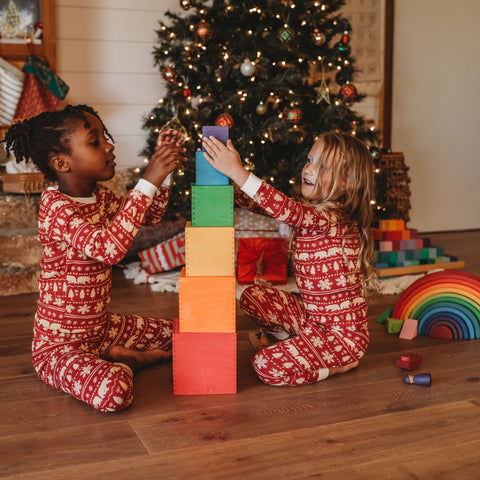 Two children sit in front of a Christmas tree, stacking wooden Grimm's rainbow boxes next to a stacking rainbow