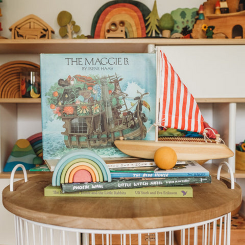 Stack of books with The Maggie B on top, a wooden toy sailboat with red and white stripped sail sits next to the book