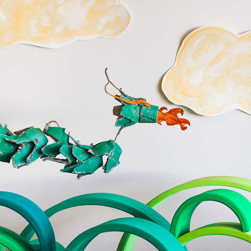 A cardboard dragon is flying over green wooden rainbows.