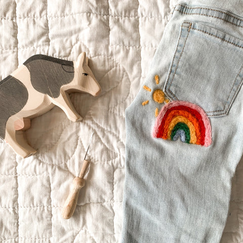 a pair of childrens jeans with a sunshine and a rainbow needle felted patch just under the back pocket sitting on a quilt next to an ostheimer cow