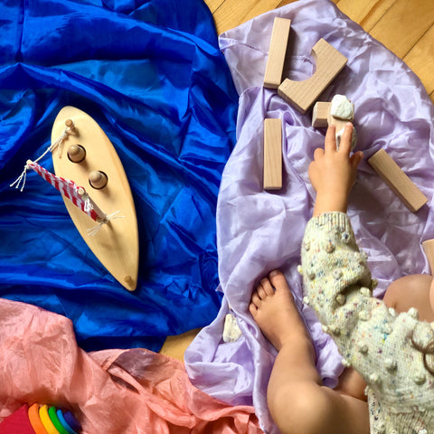 A toy wooden boat sits on a blue play silk next to a lilac play silk with a few wooden blocks on top. A child's hand reaches for the blocks. A small wooden Waldorf rainbow sits in the bottom left corner on top of a pink play silk.