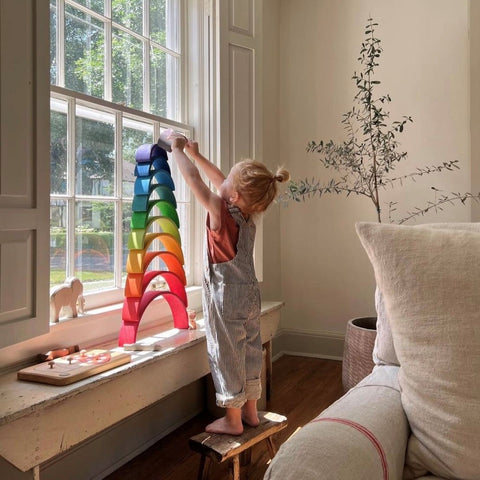 Small child in overalls reaching to the top of a stacked Grimm's wooden 12 piece rainbow in front of a window. It is stacked in rainbow order with red on the bottom and violet on the top.