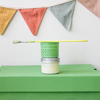 Two paint jars sit on top of a green shoebox with a paintbrush.