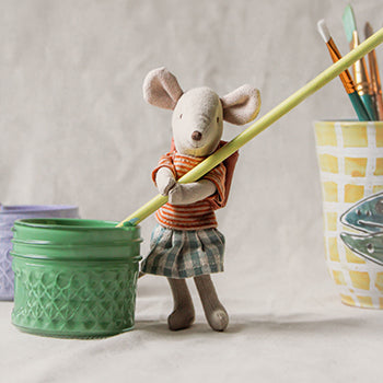 A Maileg mouse is holding a paint brush in a pot of green paint.